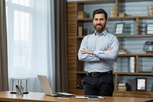 Portrait of successful businessman in office, man in shirt smiling and looking at camera, mature boss with beard with shaggy hands standing at workplace inside building. photo