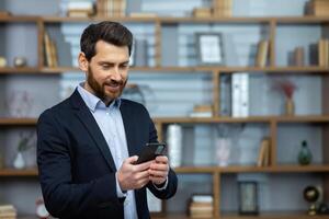 Successful mature experienced man in business suit standing near window inside office, boss using phone, typing message and browsing online pages, businessman satisfied with work. photo