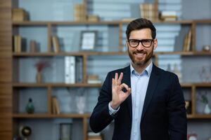 Okay firmar, empresario muestra aprobación firmar a cámara, sonriente hombre en negocio traje dentro oficina a lugar de trabajo, experimentado financiero inversor en oficina. foto