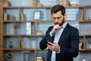 grave pensando empresario en pie cerca ventana a lugar de trabajo, mayor jefe recibido en línea notificación en teléfono con malo noticias, hombre en traje leyendo decepcionado, utilizando aplicación en teléfono inteligente foto