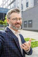 Portrait of a friendly senior businessman with a confident smile, gesturing as if to converse, standing outside an urban office building. photo