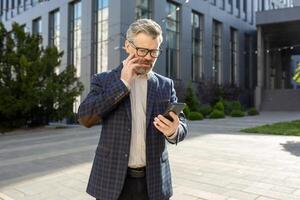 maduro masculino Finanzas experto en negocio traje comprometido en teléfono conversación, participación teléfono inteligente fuera de un oficina complejo. foto
