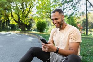 Happy and satisfied man after jogging and fitness class sitting and using smartphone, African American man in headphones listening to online podcasts, audio books and music, typing on phone. photo