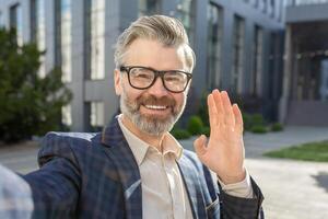 Friendly senior man in business attire waving hello, exuding confidence and professionalism outside a contemporary office building. photo