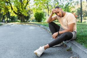 cansado africano americano hombre sentado en el suelo en el parque, hombre después trotar y haciendo activo físico ejercicio sobrecargado de trabajo descansando en un soleado día. foto