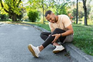 corredor lesionado su pierna mientras trotar en el parque cerca el árboles, Hispano hombre tiene músculo espasmo masajear su rodilla, hombre es corriendo y hacer ejercicio aptitud en un soleado día. foto