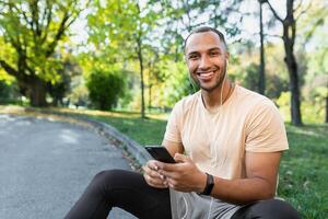 un joven afroamericano masculino deportista, atleta, corredor en auriculares se sienta en el parque en el frenar. sostiene el teléfono en su manos, escucha a música, vocación, descansar. él mira a el cámara, sonrisas foto