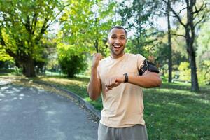 Portrait successful sportsman man satisfied with training results and achievements ,looking at camera and holding hands up in triumph gesture ,hispanic man running in park with headphones and phone. photo