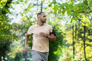 Hispanic man jogging in the park on a sunny day, runner listening to music in wired headphones, audio books and podcasts, sportsman happy with an active weekend. photo