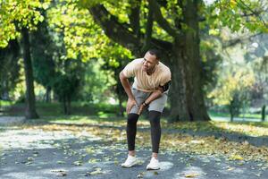 Runner injured his leg while jogging in the park near the trees, hispanic man has muscle spasm massaging his knee, man is running and exercising fitness on a sunny day. photo