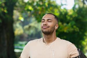 Close-up portrait of sportsman in park, hispanic man jogging in park with eyes closed breathing fresh air and resting, jogging with headphones listening to music and online radio and podcasts. photo