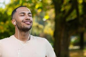 Close-up portrait of sportsman in park, hispanic man jogging in park with eyes closed breathing fresh air and resting, jogging with headphones listening to music and online radio and podcasts. photo