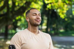 Close-up portrait of sportsman in park, hispanic man jogging in park with eyes closed breathing fresh air and resting, jogging with headphones listening to music and online radio and podcasts. photo