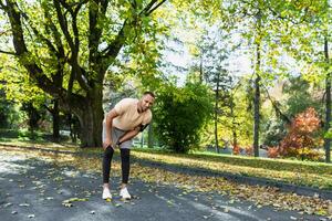 corredor lesionado su pierna mientras trotar en el parque cerca el árboles, Hispano hombre tiene músculo espasmo masajear su rodilla, hombre es corriendo y hacer ejercicio aptitud en un soleado día. foto