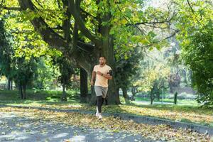 Hispanic man jogging in the park on a sunny day, runner listening to music in wired headphones, audio books and podcasts, sportsman happy with an active weekend. photo