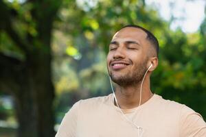 Close-up portrait of sportsman in park, hispanic man jogging in park with eyes closed breathing fresh air and resting, jogging with headphones listening to music and online radio and podcasts. photo