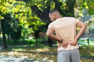 Tired man after jogging in park, hispanic man has severe pain in back and muscles after fitness, massaging his side with hand near trees. photo