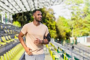 Young hispanic man with headphones running in stadium in the morning, male listening to music and audio books online podcasts using app on phone, runner smiling and happy with daily training. photo