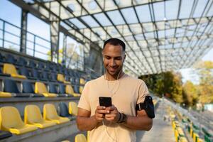 Male athlete uses app on phone after fitness class at stadium, trainer in headphones uses smartphone to listen to music, walks, smiles and rests. photo