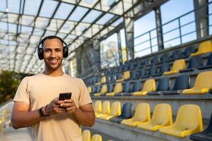 deportista utilizando teléfono sonriente y mirando a cámara, hombre en Mañana empujoncito y hacer ejercicio a Deportes estadio, hombre escuchando a música en auriculares y podcasts en línea radio. foto