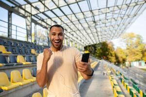 A successful man, satisfied with the result of training, looks at the camera and smiles, Hispanic holds a phone in hands uses an online sports program, an athlete holds his hand up in achievement. photo