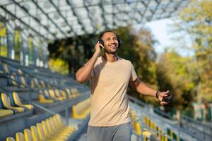 Young man walking in stadium after exercise and jogging in headphones, listening to audiobook music and online radio podcast, dancing joyfully, hispanic man on jogging day. photo