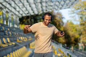 Young man walking in stadium after exercise and jogging in headphones, listening to audiobook music and online radio podcast, dancing joyfully, hispanic man on jogging day. photo