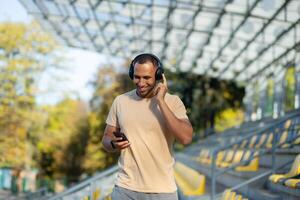 contento y sonriente indio hombre en parque estadio caminando después activo trotar y aptitud clases, utilizando teléfono y solicitud para escuchando a en línea radio podcasts y música, deportista en auriculares. foto