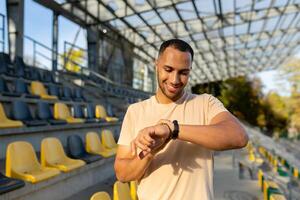 Young Latin American male sportsman stands at the stadium and looks at the fitness bracelet, the watch on his hand. Checks the time, the result, detects the stopwatch, checks the pulse, breathing. photo