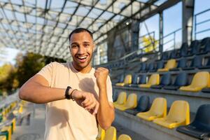 joven africano americano masculino atleta al aire libre es contento con el resultado en el aptitud pulsera. muestra un victoria gesto con un si mano, celebra, sonrisas y mira a el cámara. foto