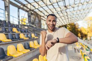 Happy young sportsman looking at camera and smiling happy with good fitness result, man checking time on fitness bracelet smart watch, running in sports stadium in daytime. photo