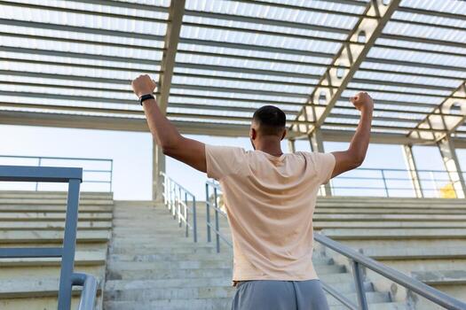 Latin american male athlete happy with training results celebrating successful sport achievement, runner standing with back near stairs holding hands up winner gesture. photo