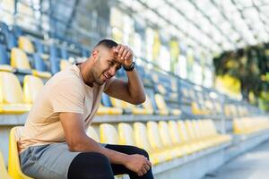 cansado y trastornado deportista atleta sentado en el estadio, africano americano hombre insatisfecho con el resultado de capacitación, hombre descansando después activo físico ejercicios y aptitud física. foto