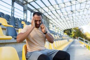 Head injury in sports. Young hispanic male postman sits in the stadium and holds his head, feels severe pain, pressure, calls an ambulance, doctor, clinic for help. photo