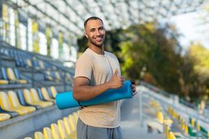 Portrait of hispanic male fitness trainer at sports stadium, athlete with yoga mat smiling and looking at camera, athlete in sportswear at sunset showing thumbs up. photo