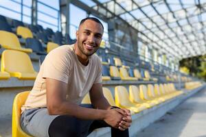 retrato de joven atleta en estadio, hombre sentado en silla sonriente y mirando a cámara cerca arriba, corredor descansando desde formación y aptitud física. foto