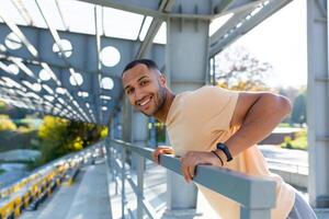 Close-up photo. Portrait of a young African American sports man standing in the stadium, doing push-ups on the bar, swinging his arms, looking at the camera smiling. photo