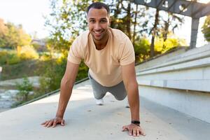 Cheerful and smiling hispanic man doing outdoor exercise in stadium, man stretching and recuperating before jogging in summer park. photo