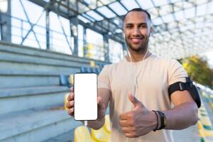 Young hispanic sportsman smiling and looking at camera, man in stadium showing white phone screen, while jogging and doing active exercise. photo