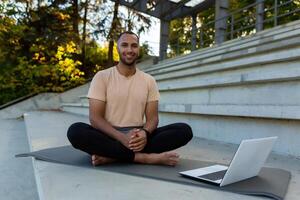 Portrait of arab sportsman fitness trainer, man smiling and looking at camera sitting on sports mat in stadium, using laptop to teach online course photo