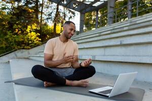 Young African American athlete fitness trainer conducts online training sitting in lotus position, man talking to physical class students using laptop and online call. photo