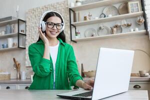 A young woman in headphones sits at home in the kitchen in front of a laptop and records a podcast, blog, growth and development training. Narrates the course, the lecture. Smiling at the camera. photo