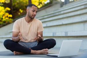 Young African American athlete fitness trainer conducts online training sitting in lotus position, man talking to physical class students using laptop and online call. photo