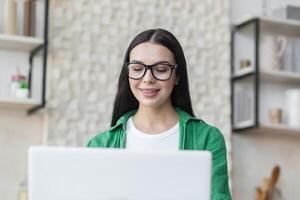 Close-up photo. A beautiful young woman is sitting at home in the kitchen using a laptop and writing and blogging, social networking, and texting with friends. photo