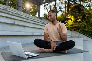 A young Latin American male sportsman, instructor, trainer sits in a stadium on a mat in front of a laptop and teaches online training. photo