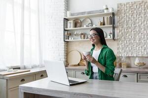 un joven mujer se sienta a hogar en el cocina con un ordenador portátil y bebidas Mañana café, participación un taza en su manos. café descanso durante trabajando horas. descansando, mirando soñando fuera el ventana, sonriente. foto