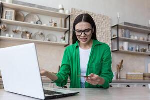 Worried young woman sitting at home in kitchen in front of laptop and looking shocked at credit card. Inexplicably spreads his hands. Financial problems, bankruptcy, blocking. photo