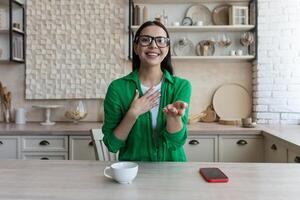 A young female blogger conducts a remote online broadcast, talks to the camera from home. Sitting in the kitchen and recording a , greetings. smiles, presents. photo