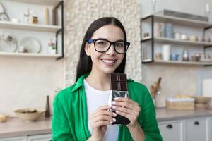 retrato de un hermosa sonriente joven mujer en pie a hogar en el cocina, participación y comiendo un chocolate bar en un envoltura. sabores, posa, mira dentro el cámara. foto