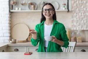 A young woman sitting in the kitchen with a cup of coffee in front of the camera and vlogging, recording a blog, conducting a webinar, growth and development training, pointing at the camera, smiling. photo